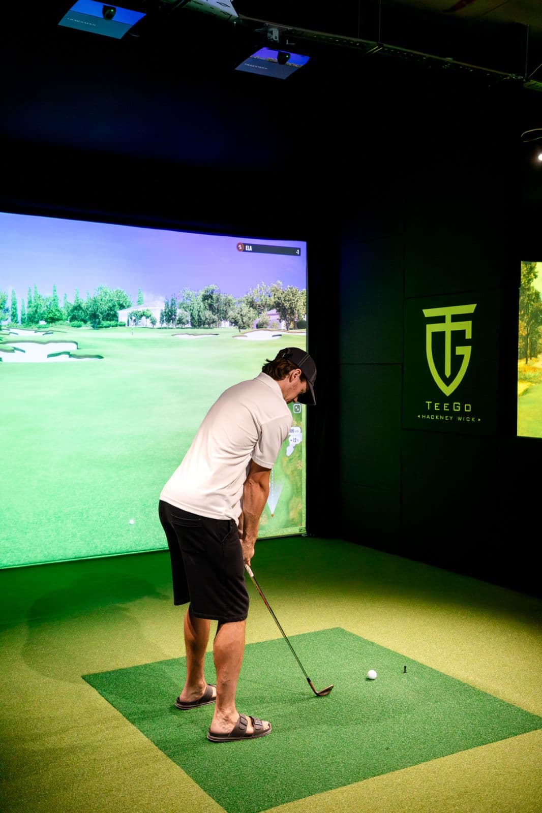 A player addressing the ball in a TeeGo simulator bay. The impact screen fills the end of the bay, hanging flat and taut with the projected course sharp across it.