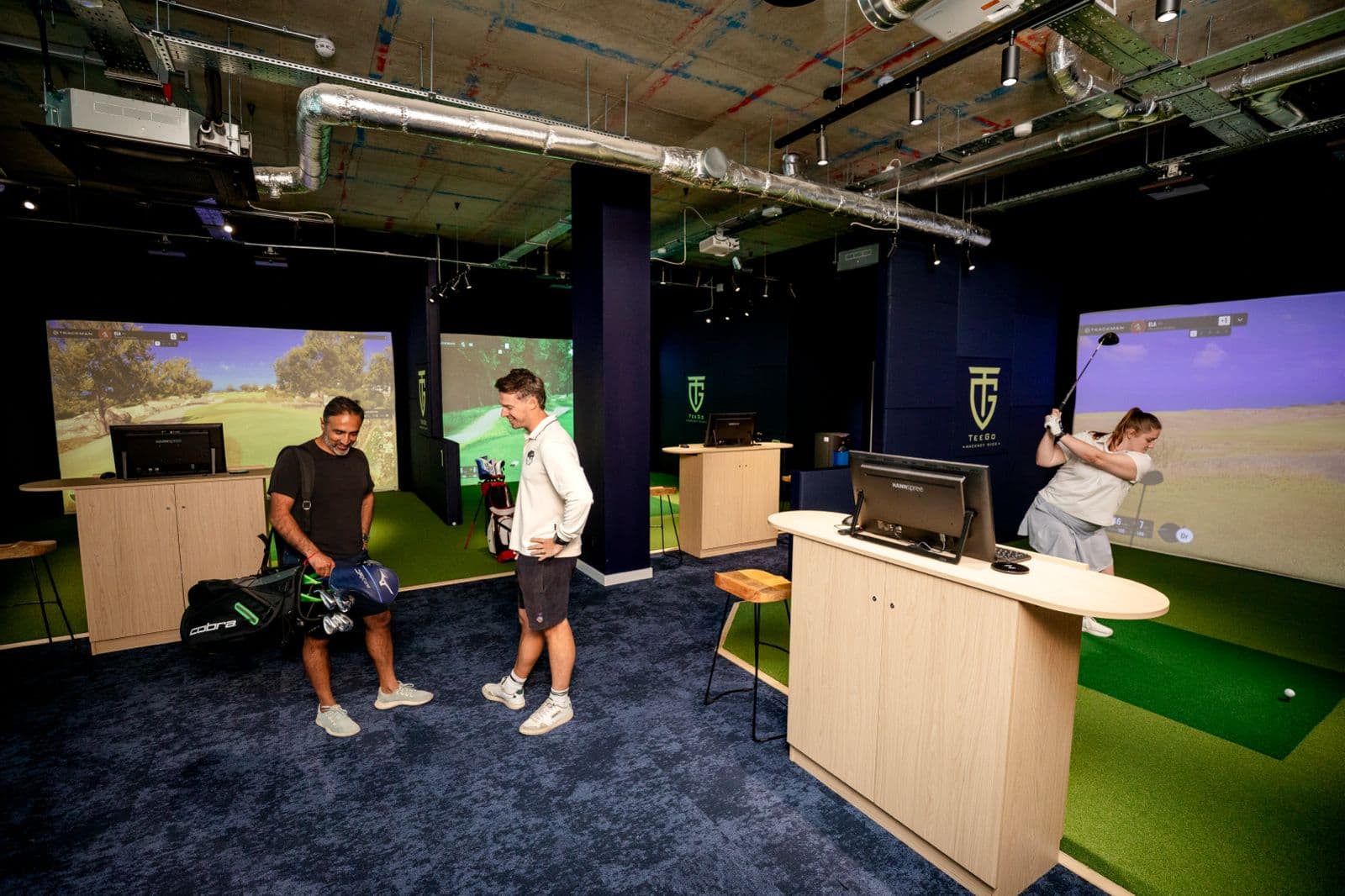Three golf simulator bays at TeeGo Hackney Wick, each with an impact screen showing a projected course. Players are mid-round; the venue has exposed services overhead and TeeGo branding on the walls.